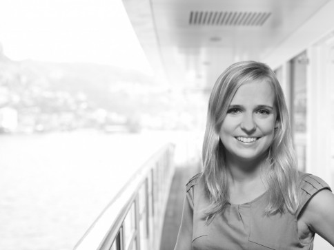 A young woman with long blonde hair smiles at the camera whilst standing on the deck of a boat. The background shows a blurred view of water and distant buildings. The image is in black and white.