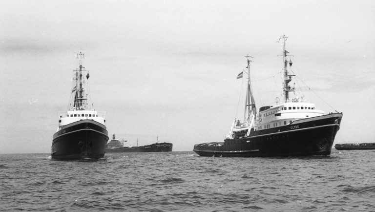 Two large tugboats are positioned on the water, one facing forward and the other angled slightly to the side, with another large ship visible in the distant background under an overcast sky.