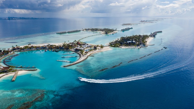 Aerial view of tropical islands surrounded by turquoise sea, with overwater bungalows, sandy beaches, and a speedboat leaving a white wake across the water under a partly cloudy sky.