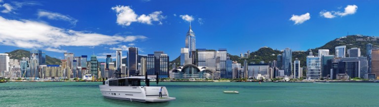 A modern boat floats on turquoise water in front of a city skyline with tall skyscrapers, green hills, and a bright blue sky with scattered clouds.