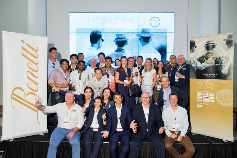 A large group of people pose together indoors at The Superyacht World Club event, smiling and holding drinks. Banners for Benetti and the event are displayed on either side, with a screen in the background.