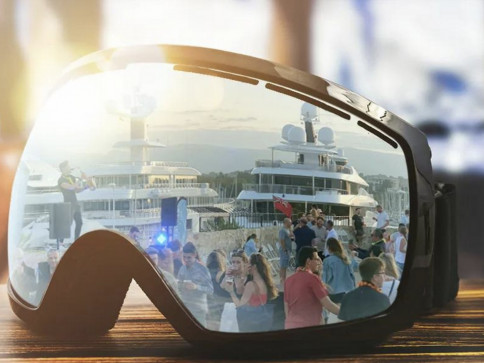 Ski goggles reflect a lively marina scene with people socialising near luxury yachts under bright sunlight, capturing a festive atmosphere by the water.
