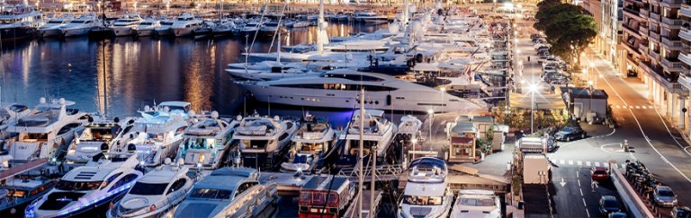 A marina at dusk filled with numerous luxury yachts and boats, with city lights reflecting on the water and surrounding buildings, creating a lively and elegant waterfront scene.