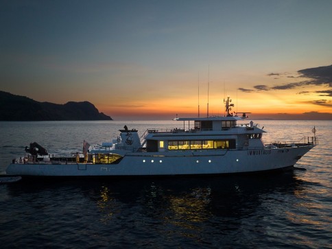 A large white yacht is anchored on calm water at sunset, with a colourful sky and a silhouette of a mountainous coastline in the background. The yacht is illuminated, highlighting its sleek, modern design.