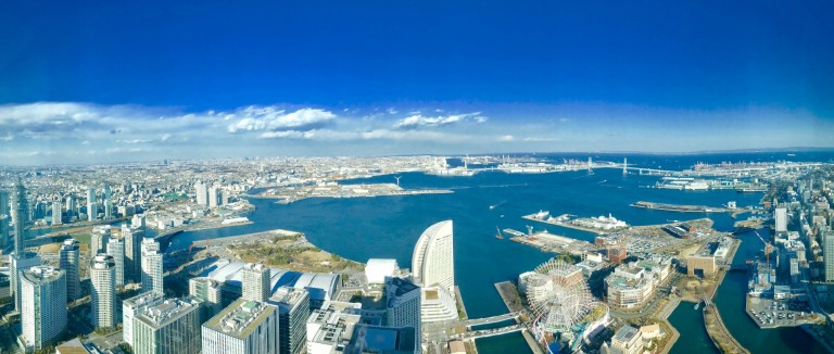 A panoramic view of a coastal city with high-rise buildings, a large bay, bridges, and clear blue sky, taken from a high vantage point during the daytime.