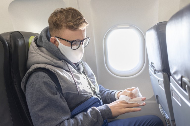 A person wearing glasses and a face mask is seated on an aeroplane, cleaning their hands with a sanitising wipe. Sunlight shines through the window beside them.