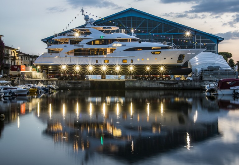 A large yacht with lights on is moored near a marina at dusk, reflecting on the still water. The background features a large, blue-roofed building and a partly cloudy sky.