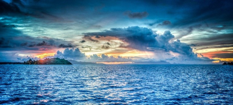 A vibrant ocean scene at sunset with choppy blue water, a small lush island on the left, and dramatic clouds in a colourful sky of blues, oranges, and purples.