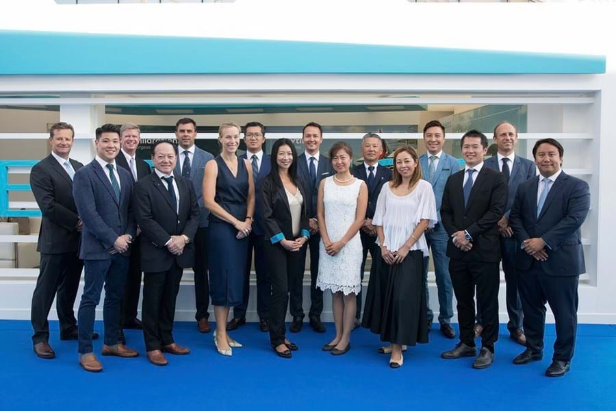 A group of seventeen people, men and women in business attire, stand and smile for a group photo on a blue carpet in front of a modern white and blue background.