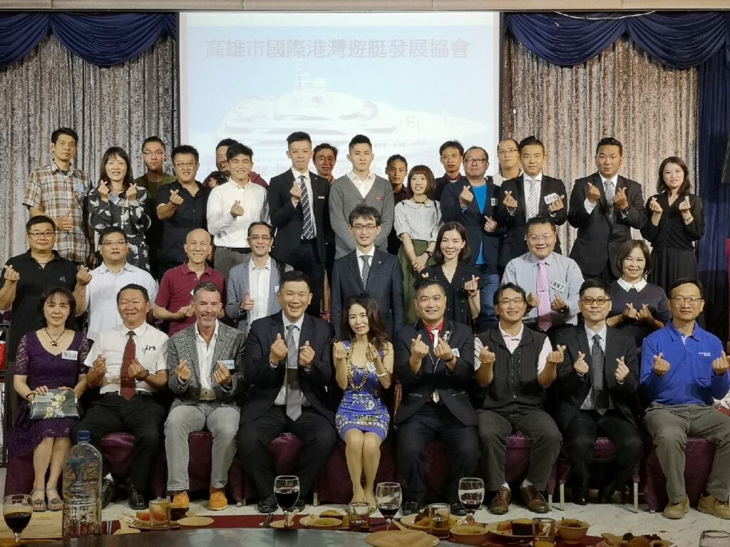 A large group of people pose indoors for a formal group photo, many making finger heart gestures. They are dressed in business or semi-formal attire, seated and standing in rows, with a presentation screen behind them.