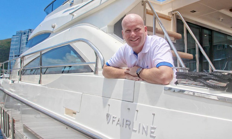 A smiling man in a white shirt leans on the rail of a moored luxury yacht named Fairline, with modern buildings and hills in the background.