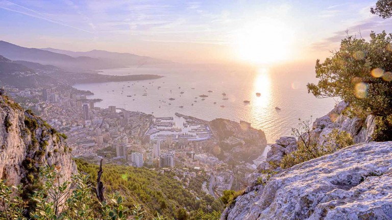 A scenic view of Monaco at sunset, with sunlight reflecting on the sea, numerous yachts in the harbour, and city buildings nestled along the coastline and surrounding hills. Rocky cliffs and greenery frame the foreground.