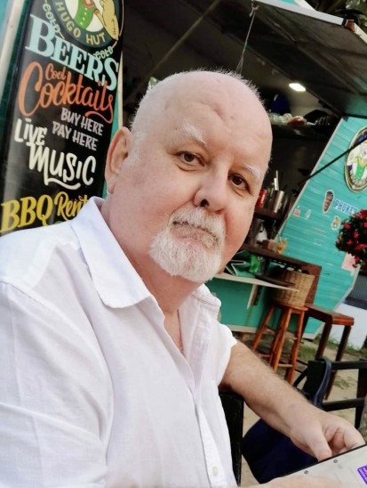 An older man with a white beard and moustache, wearing a white shirt, sits outdoors at a restaurant; a sign behind him advertises beers, cocktails, and live music.