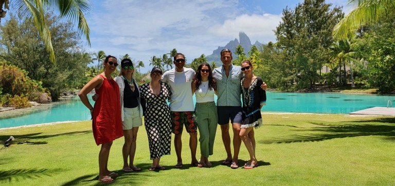 A group of seven people stand together smiling on a grassy lawn in front of a turquoise lagoon with palm trees and mountains in the background on a sunny day.