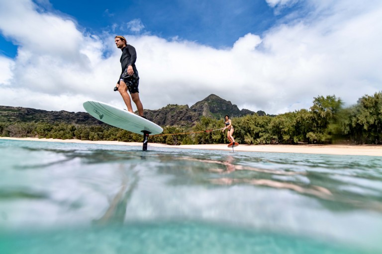 A man rides a hydrofoil surfboard above the water whilst a woman balances on a slackline stretched over the shallow sea near a sandy beach with lush green mountains in the background.