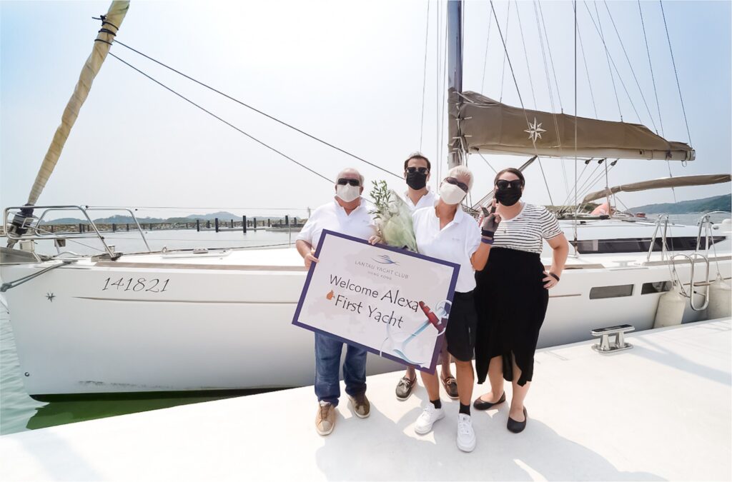 Four people wearing masks stand on a quay in front of a sailing boat, holding a sign that says Welcome Alex! #FirstYacht and a small plant. The weather is sunny and clear, and the water and boat are visible in the background.