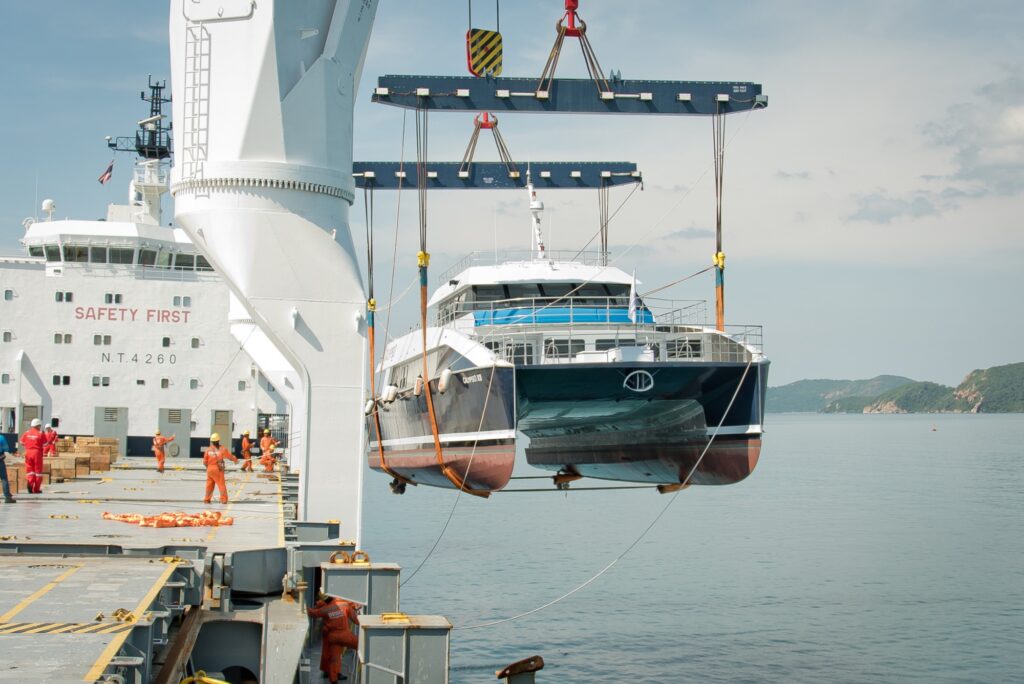 A large ship uses a crane to lift a blue and white catamaran from the water. Several crew members in orange suits stand on the deck. Calm water and distant hills are visible in the background.