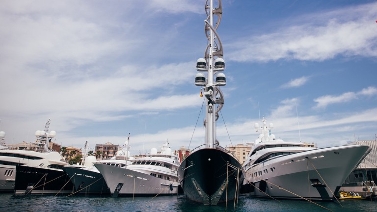 Several large luxury yachts are moored side by side in a marina under a partly cloudy sky, with buildings visible in the background. The central yacht features a distinctive, intricate mast design.