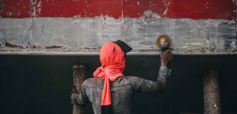 A person wearing a red headscarf and dark clothing is sanding or polishing a weathered metal surface with a power tool, facing away from the camera.