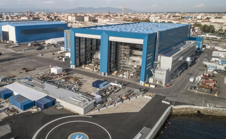 A large blue industrial building with open sections revealing construction inside, surrounded by storage containers and equipment, located near a waterfront with a helipad and cityscape in the background.