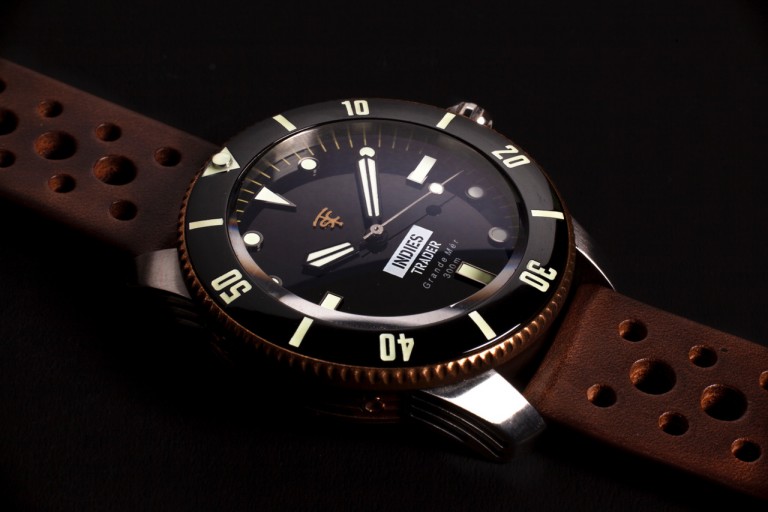 Close-up of a wristwatch with a brown perforated leather strap, black dial, luminous hour markers, and rotating bezel, displaying the day and date. The background is dark, highlighting the watch’s details.