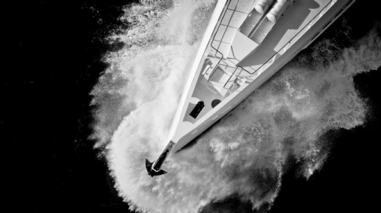 Aerial view of a boat cutting through water at high speed, creating a dramatic splash and waves, seen in black and white.