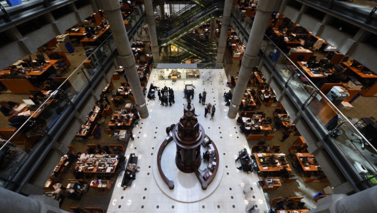A large, open-plan office with multiple desks and people working, viewed from above. The central area features a tall, ornate structure, surrounded by groups of people and modern industrial architecture.
