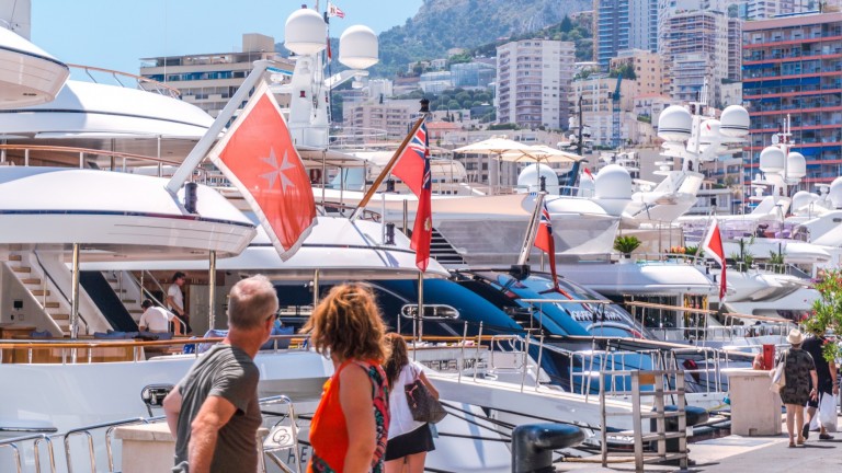 Two people walk along a quay lined with luxury yachts displaying red maritime flags, with tall buildings and a hillside in the background on a sunny day.