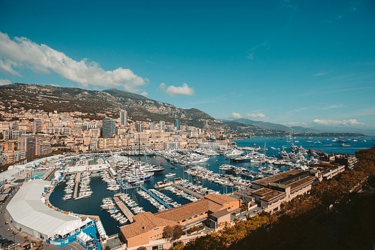 A panoramic view of a marina filled with yachts and boats, surrounded by buildings and mountains under a blue sky in a coastal city.