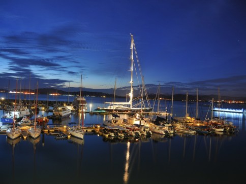 A marina at dusk with several boats and yachts moored on calm water, illuminated by soft lights. The sky is dark blue with scattered clouds and distant city lights visible on the horizon.