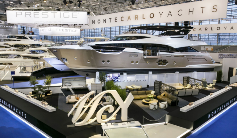 A large, luxurious yacht is displayed indoors at a boat show. The area features modern seating, decorative plants, and prominent MCY and Monte Carlo Yachts branding on banners above and around the exhibit.