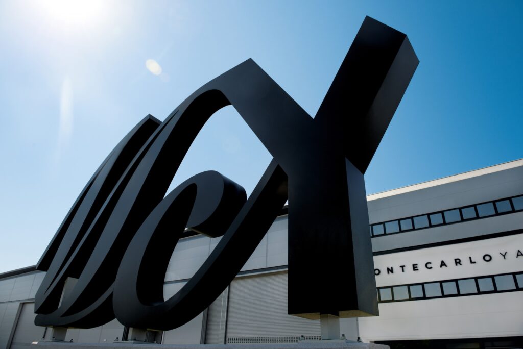 A large, black, stylised MCY sculpture stands in front of a modern white building with Montecarloyachts partially visible on the facade under a clear blue sky.