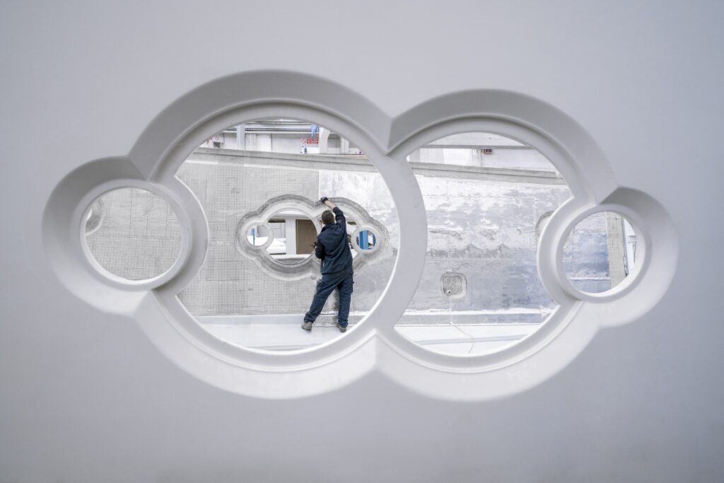 A person in dark clothing works on a large industrial structure, seen through two circular windows with decorative framing in the foreground. The scene has a clean, modern, and industrial aesthetic.