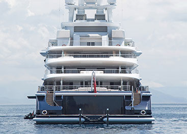 A luxury yacht viewed from the rear, showing multiple decks with railings, a small boat alongside, and a swim platform extending into the calm blue water. The sky is lightly clouded in the background.