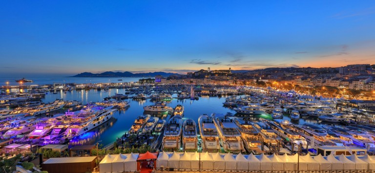 A vibrant marina at sunset with numerous yachts moored in the harbour, city buildings and hills in the background, and the sky glowing with shades of blue and orange.