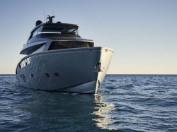 A sleek, modern yacht is seen from the front, floating on calm blue water under a clear sky at sunset.