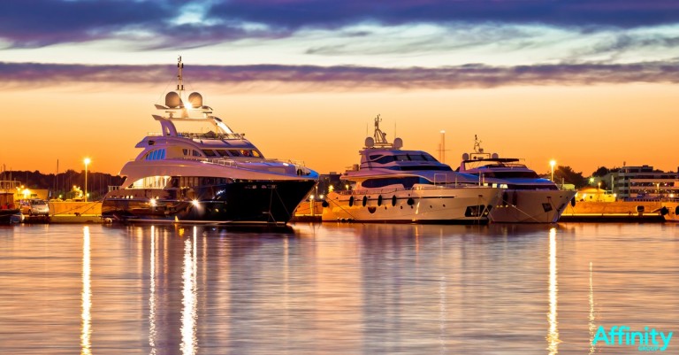 Luxury yachts moored at a marina during sunset, with colourful sky reflected on the calm water. The marina lights and silhouettes of distant buildings add to the tranquil evening scene. The word Affinity appears in the corner.