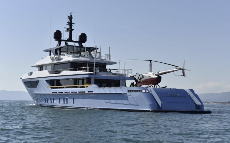 A large modern yacht with a helicopter parked on its rear deck floats on calm blue water, with mountains and a clear sky visible in the background.