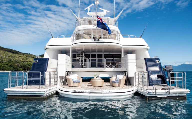 A luxurious white yacht anchored in clear blue water, featuring an open deck with modern outdoor seating, two side platforms, and a Cook Islands flag flying at the top. Lush green coastline and mountains appear in the background.