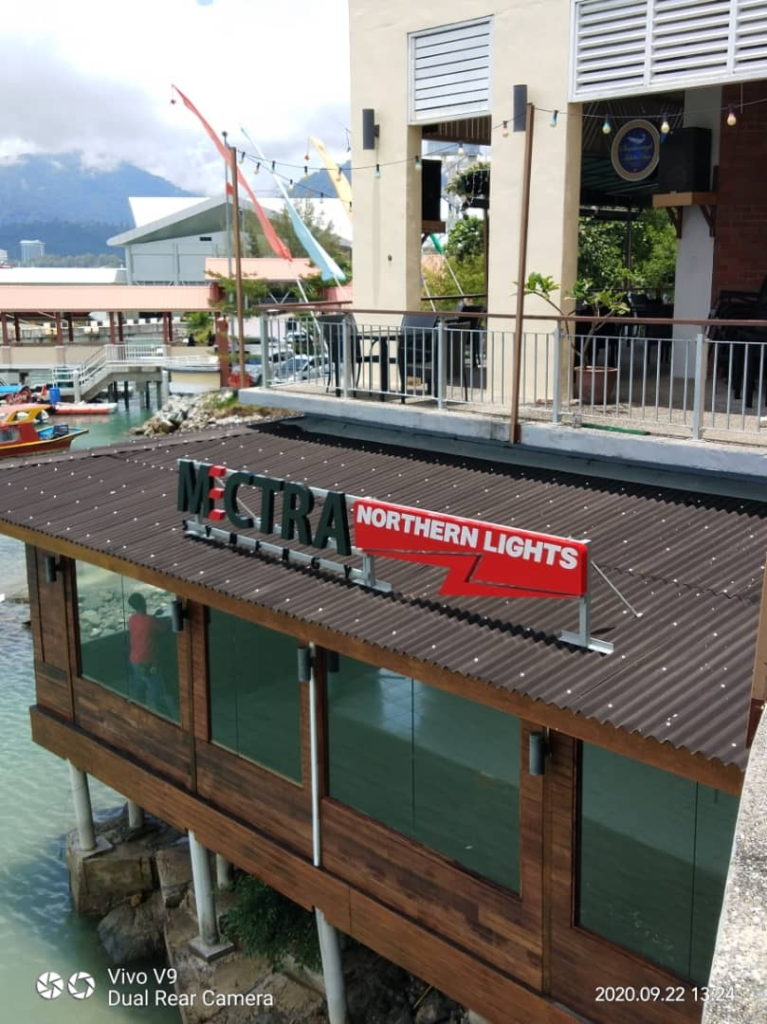A waterfront restaurant with a rooftop sign reading “MICTRA NORTHERN LIGHTS.” The building has wooden walls, large windows, and is situated by the water. Another building and boats are seen in the background.
