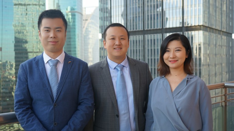 Three people in business attire stand on a balcony with tall glass office buildings in the background, smiling at the camera.