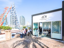 People gather outside a modern waterfront building with a sign reading ICV under a clear blue sky. Tall buildings and sail-shaped sculptures are visible in the background.