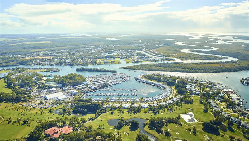 Aerial view of a marina filled with boats, surrounded by green golf courses, residential areas, winding rivers, and lush forested landscape under a partly cloudy sky.