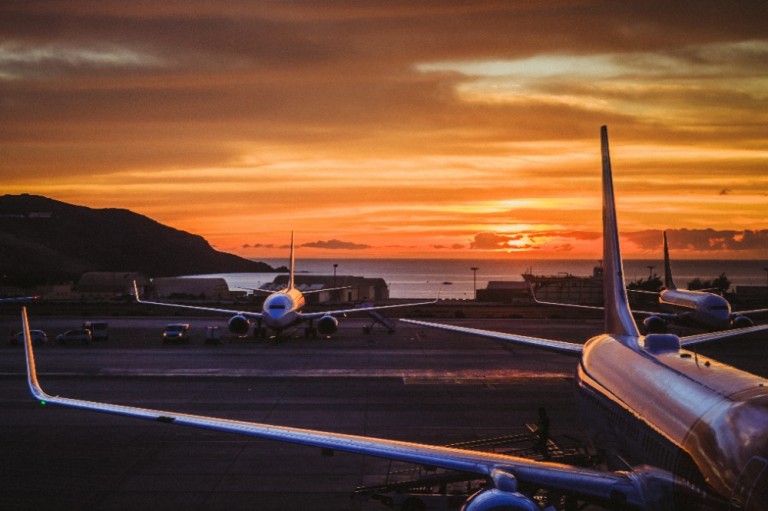 Three aeroplanes are parked on an airport runway at sunset, with a dramatic orange and yellow sky and a mountain in the background. The sun is low on the horizon, creating reflections on the aeroplanes.
