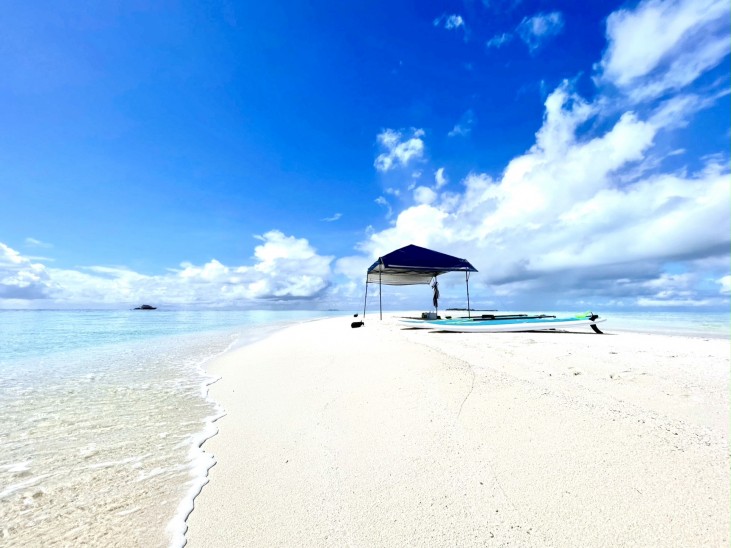 A small canopy stands on a pristine white sandy beach surrounded by clear blue water under a bright blue sky with scattered clouds. A paddleboard rests next to the canopy.