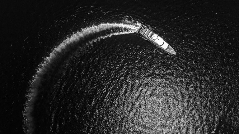 Aerial view of a yacht making a sharp turn on dark water, leaving a curved white wake behind it. The sunlight reflects off the rippled surface of the water.